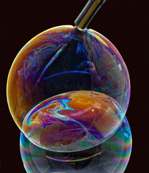photo of two bubbles being blown out of a straw, their vivid swirling rainbow colors stand out against the black background